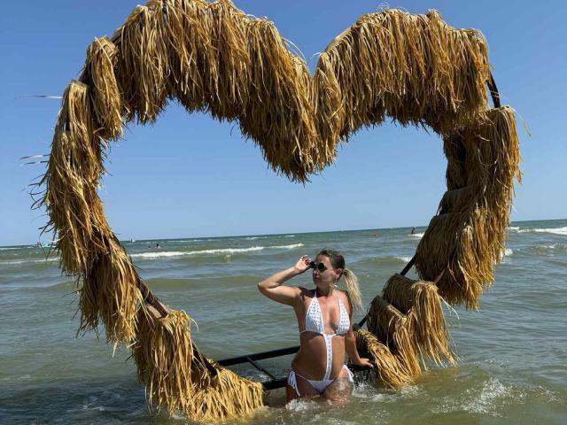 AngryGirl69 in a white bikini stands in shallow water, hands on head, in front of a heart-shaped straw frame. She has blonde hair and sunglasses.