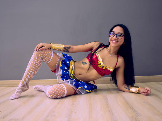 SofiaaMiiller, wearing a Wonder Woman costume, has small tits, a red bra, blue skirt, white fishnet stockings, and glasses, lying on wooden floor, smiling, with tattoos visible.