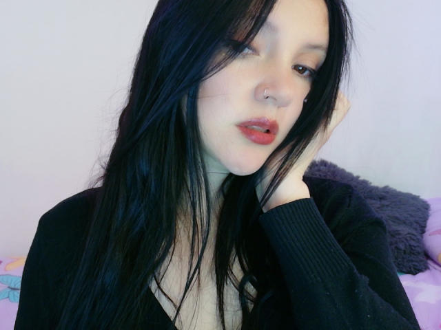 NathalyCurvy with black hair, nose ring, and red lipstick, touching her face with a black cardigan, showing a bit of her cleavage, looking slightly bored or pensive.