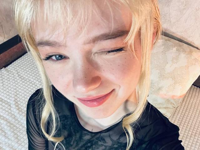 EnchantingEyes, a blonde with wavy hair, is winking with one eye, showing off her cute freckled face, full pink lips, and black lace top, sitting on a white bed.