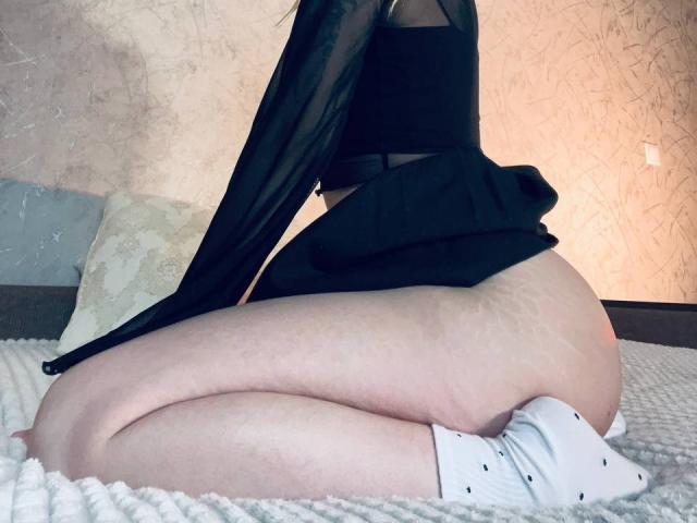 EnchantingEyes is kneeling on a bed, wearing a black sheer top and no panties, exposing her pale, stretch-marked ass and white socks with black dots.