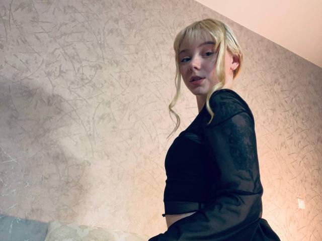 EnchantingEyes with blonde hair, wearing a black sheer top, looking over her shoulder, showing her small tits and a subtle ass, in a dimly lit room.