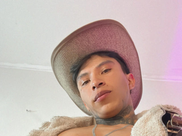 MickeFire has a tan face, short black hair, and tattoos on his neck, wearing a gray hat and a fluffy beige jacket, with his tongue slightly out.