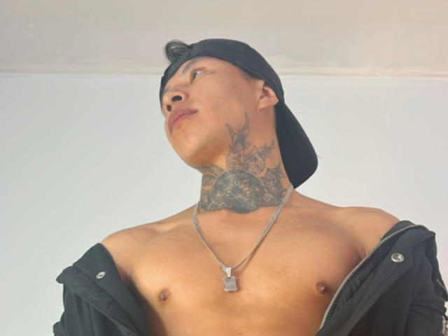 MickeFire, shirtless with black jacket off, wearing black cap, looking up, tattoos on neck, chest, and necklace with dog tag. Light skin, tattoos, cap, necklace.