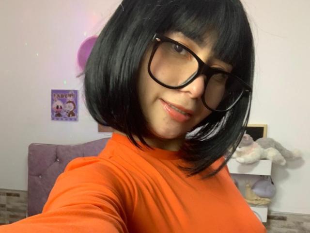 MelannyTaylorr com cabelo curto preto, óculos, blusa laranja, sorrindo para a câmera, mostrando os seios, rosto fofo, pele média, quarto branco, cabeceira roxa, brinquedo de pelúcia à direita..