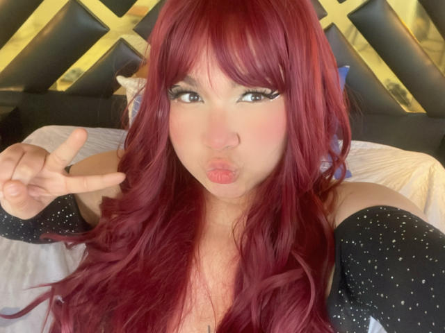 LillyMayer with red hair, pouty lips, and a peace sign, wearing a sparkly black top, has big tits and is making a cute face.