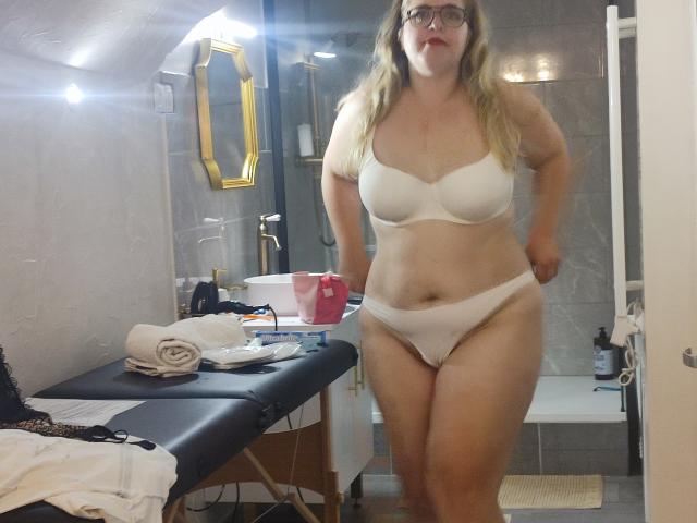 Bonnefrancaise stands in a bathroom wearing white lingerie, with her hands on her hips, showing her tits and pussy. She has glasses and blonde hair, looking at the camera.