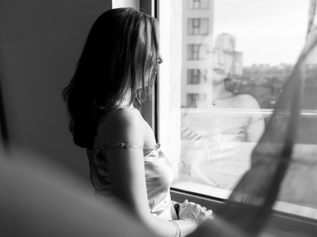 Black-and-white photo of AmethystCharm with medium tits, wearing a silky off-shoulder top, standing by a window, gazing outside, with a curtain on the right.