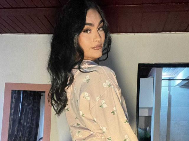 KamilaBabe with long black hair, wearing a pink floral shirt, looking over her shoulder, showing her ass, in a dimly lit room with a mirror.