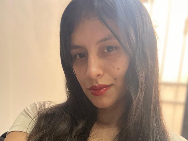 MadissonFontaine with long black hair, red lipstick, freckles, and a small mole on her left cheek, wearing a white shirt, smiling softly at the camera.