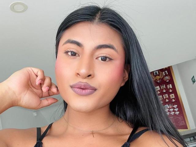 MichelleDenise has tan skin, black straight hair, brown eyes, pink lipstick, and a cute face. She's wearing a black top and a thin necklace, with her hand near her face.