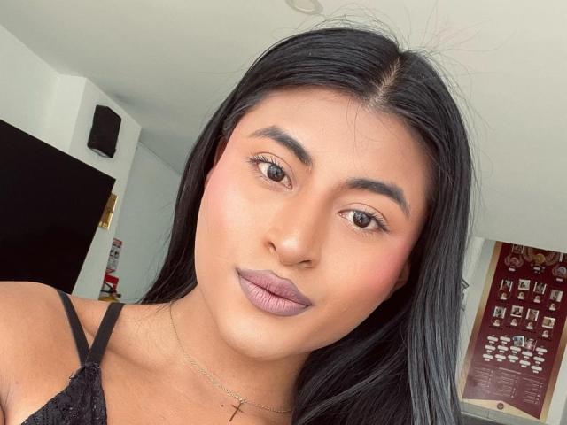 MichelleDenise has smooth brown skin, long black hair, and purple lipstick. She's wearing a black lace top, with a cross necklace. Her tits are small, and she's looking at the camera.