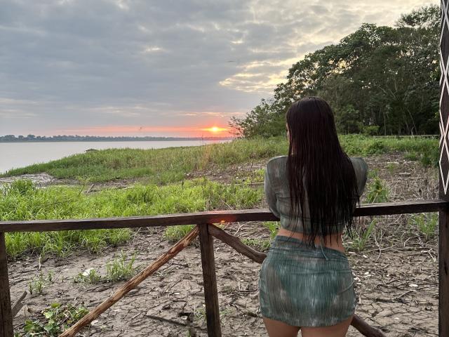 VictoriaWaynes with long dark hair, facing sunset, wearing a gray crop top and denim skirt, standing on a wooden railing, green grass, and trees in the background.