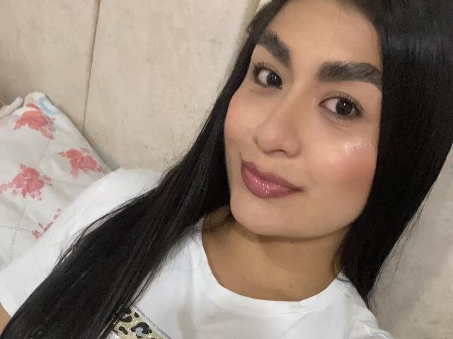 MiaDuboisX ha lunghi capelli neri, occhi marroni e un rossetto rosa lucido. Indossa una camicia bianca con stampa leopardata e siede su un divano beige..