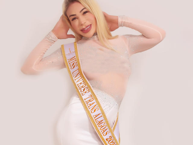 TatuadaSafadaHot mit blonden Haaren, die ein durchsichtiges, glitzerndes Oberteil und weiße Hosen trägt, mit einem Band mit der Aufschrift "Miss Thailand International" über ihrer Brust, lächelnd, mit den Händen hinter dem Kopf..