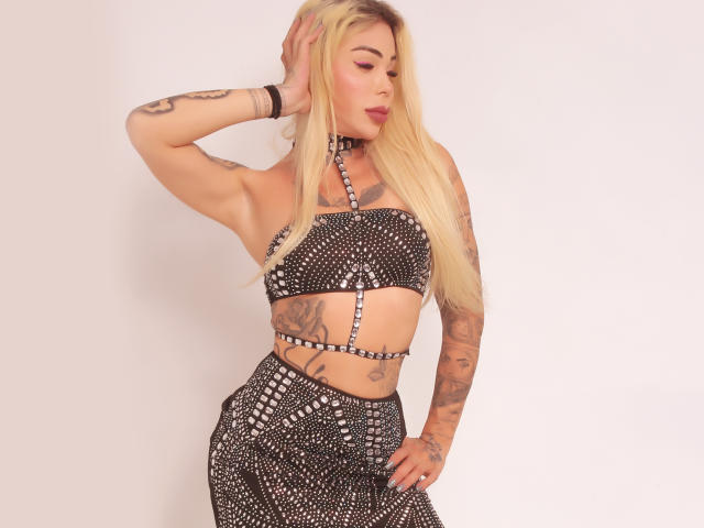 TatuataSafadaHot con lunghi capelli biondi, tatuaggi, che indossa un top e una gonna neri con borchie, una mano tra i capelli, che guarda di lato, mostrando il seno e i glutei..