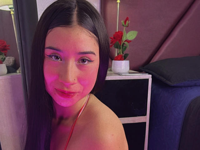 SweetyLuna with pink-tinged face, long brown hair, and red lipstick, wearing a red necklace, standing near a black bed with a red rose in a white pot.