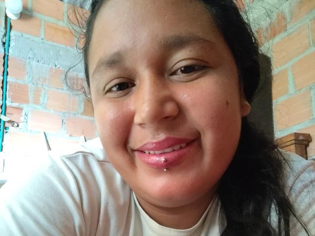 SamiBeauty has a chubby face, brown eyes, black hair, and a white shirt. She's smiling with a pierced bottom lip and a small beauty mark on her right cheek.