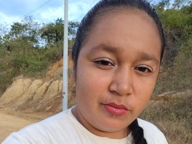 SamiBeauty's face, medium brown skin, black hair in braid, white shirt, small lip piercing, slight smile, brown eyes, soft expression, outdoor background, clear blue sky.