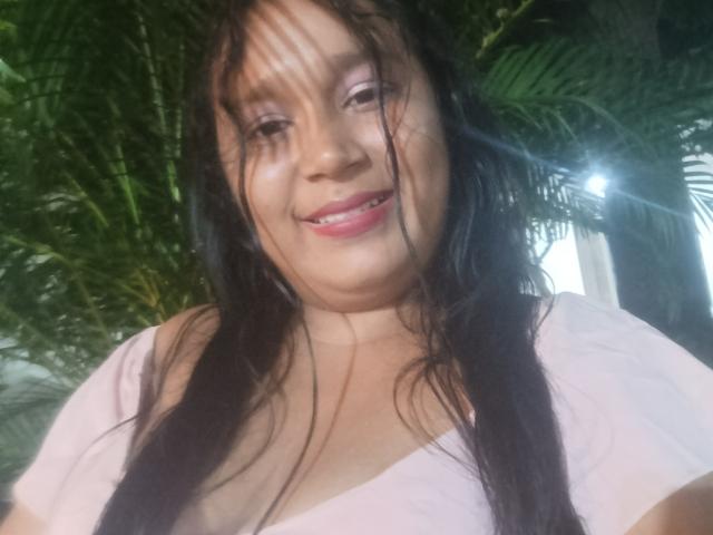 SamiBeauty has wet black hair, pink lipstick, and a cute smile. She's wearing a pink top, showing her tits slightly. Background is green with plant leaves.
