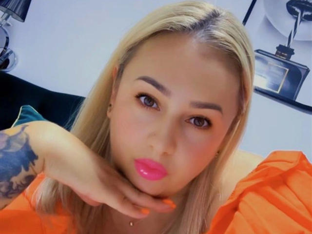 krystal75-hot with blonde hair, pink lips, orange shirt, hand on cheek, tattoo on arm, orange nails, looking at camera, brown eyes, subtle makeup, sexy.