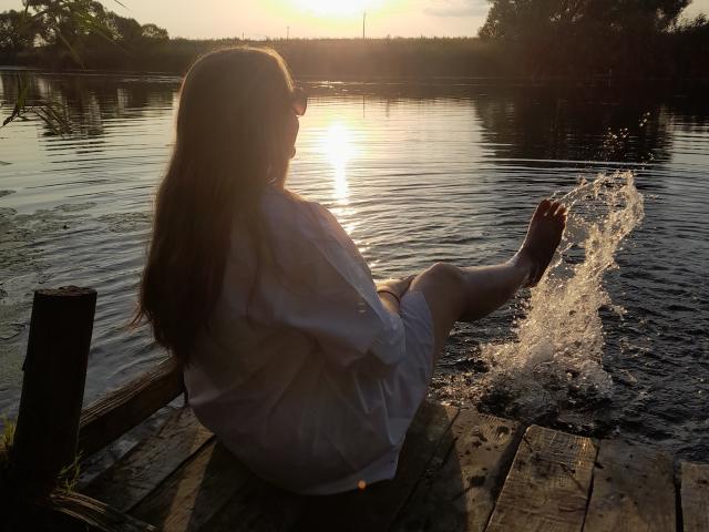 RareLazery, with long brown hair, sits on a wooden dock, splashing water with her right foot, wearing a white dress, sun setting in the background.
