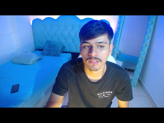 CharlieeCock, a medium-build guy with short dark hair and a mustache, wearing a black t-shirt, sitting on a bed with blue lighting, phone on the bed.