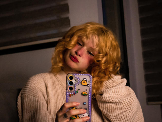 DanaStone with curly blonde hair, wearing a beige knitted sweater, taking a selfie with a phone covered in cute yellow duck stickers, has pink blush on her cheeks.