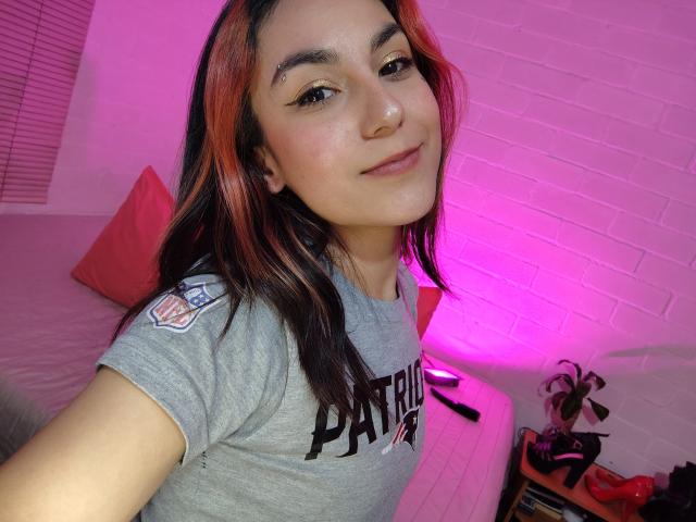 AmyStreetwise69 with red and black hair, light skin, and eyeliner, wearing a grey "Patrion" shirt, smiling slightly, pink room lighting, white brick wall, and a bed in the background.
