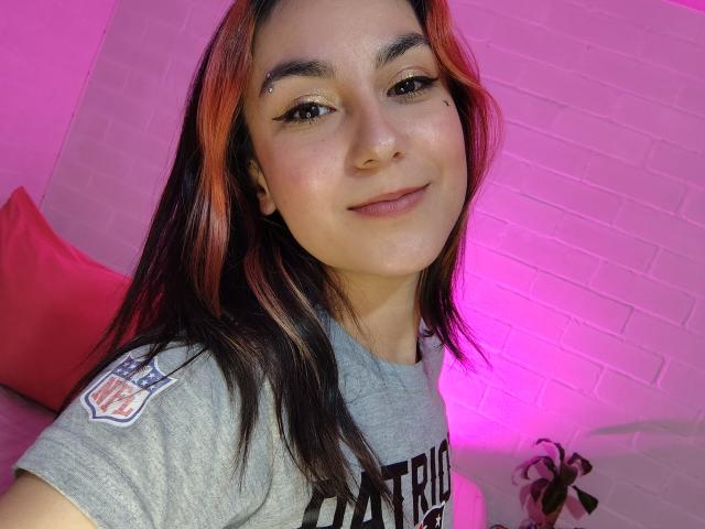 AmyStreetwise69 with long, dark brown and orange-dyed hair, wearing a grey t-shirt, has a nose ring, black eyeliner, and a subtle smile, with a pinkish-purple light.