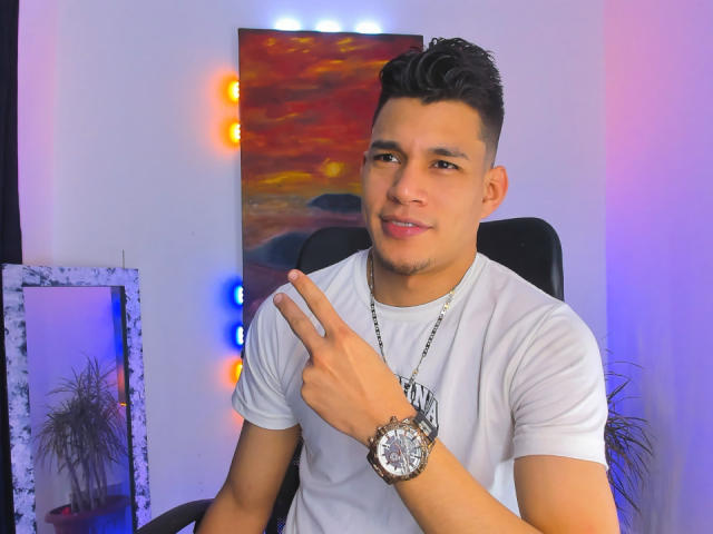 TheNewMan, ein süßer Latino-Typ mit kurzen schwarzen Haaren, einem weißen T-Shirt und einer silbernen Uhr, zeigt mit einem leichten Lächeln auf die Kamera und präsentiert dabei sein Handgelenk..