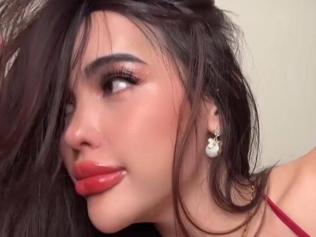 LucineDelacruz's face, with red lipstick, slightly parted glossy pink lips, dark wavy hair, and a heart-shaped earring, looking to the side with a subtle smile.