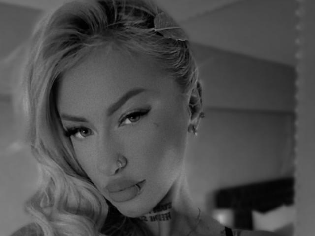 SinnersMuse with blonde hair, a nose ring, and a choker, looking seductively at the camera. Greyscale, close-up, wavy hair, eyeliner, and a slight pout.