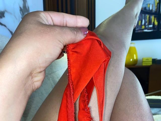 La mano de SofiaaTailor sosteniendo unas panties rojas contra su pierna desnuda, ligeramente bronceada, con vello y pecas, con esmalte de uñas rosa, en una habitación bien iluminada..
