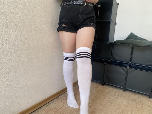 Reiza's legs in white knee-high socks with black stripes, black ripped denim shorts, and a black belt with a buckle, standing on a beige carpeted floor.