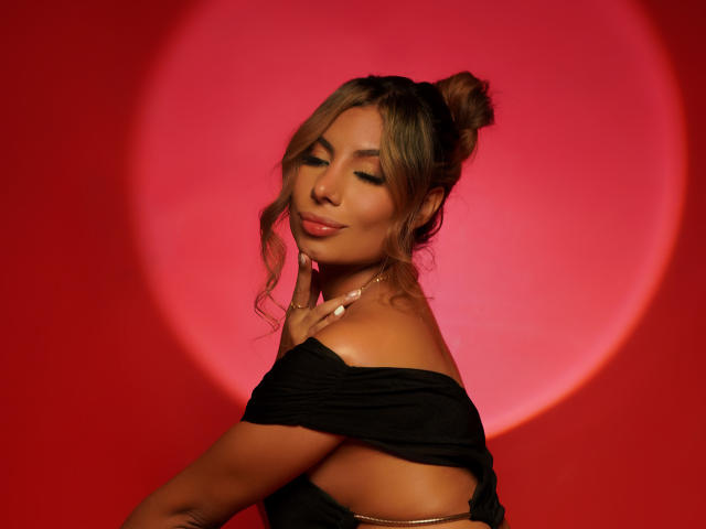 AmyBermudez with cute, wavy brown hair in a bun, smiling, eyes closed, black off-shoulder top, finger on cheek, glowing red circular light behind her, showing her tits.