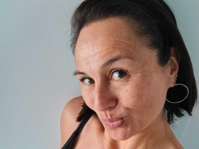 LatinLola has medium brown skin, dark brown eyes, and short black hair. She's pouting her lips, wearing black earrings, and a black strap is visible on her shoulder.