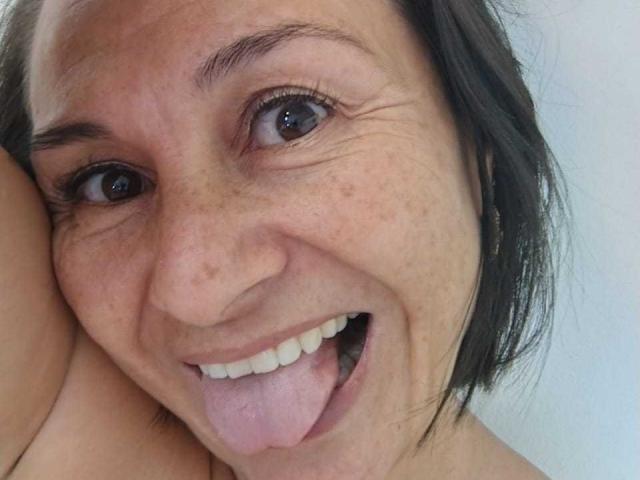 LatinLola with freckled face, dark brown eyes, short black hair, and tongue out, smiling at the camera, showing white teeth and slightly parted lips.