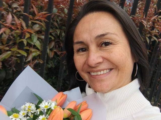 LatinLola with medium brown hair, smiling, holding a bouquet of orange tulips and white daisies, wearing a white turtleneck, and large hoop earrings.