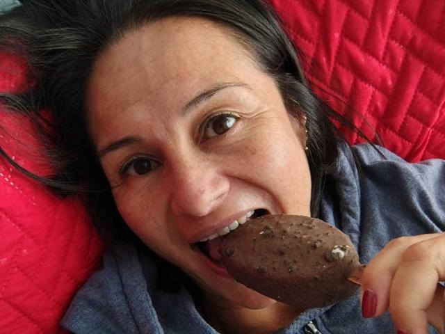 LatinLola with dark hair and brown eyes, tongue out, eating a chocolate-covered cookie, red quilted blanket in background, wearing blue hoodie, holding cookie with red-painted nails.