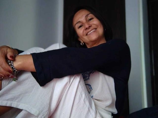 LatinLola with black hair, smiling, wearing a navy top, white skirt, and bracelets, hugging her boobs, arms crossed, showing red nails, and a happy expression.