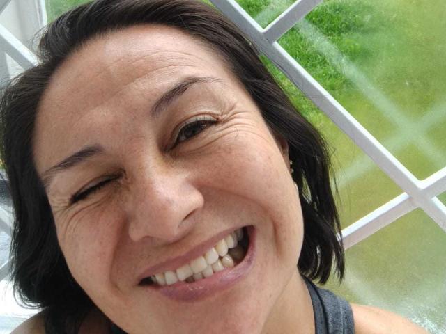 LatinLola has a big, happy smile with her eyes closed, showing her white teeth. She has dark brown hair, a freckled nose, and is wearing a gray tank top.