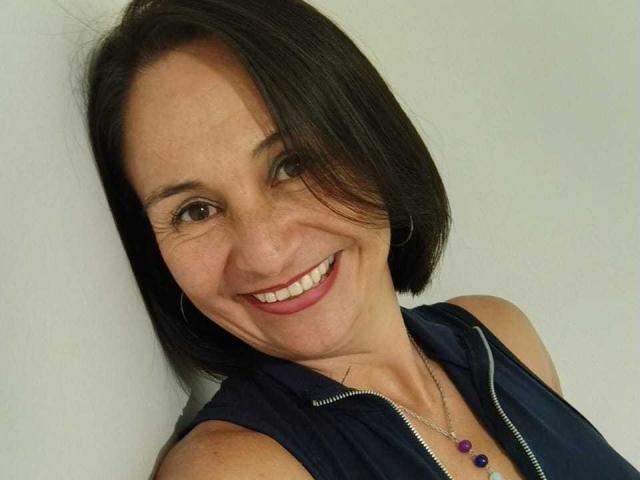 LatinLola, smiling with short black hair, wearing a sleeveless navy top with a zipper, and a necklace with purple beads, has her ass slightly visible in the image.