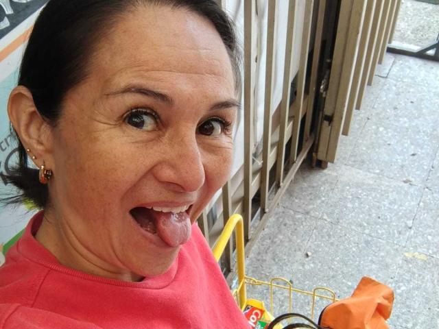 LatinLola's face, tongue out, wearing a red shirt, brown hair pulled back, earrings, yellow shopping cart with orange bag, metal fence, concrete floor, smiling, slightly wrinkled skin.