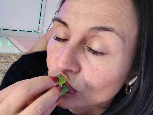 LatinLola with closed eyes, dark hair, and fair skin, eating a small green leaf with red-painted nails, wearing white earrings, looking slightly up, in a relaxed, intimate moment.