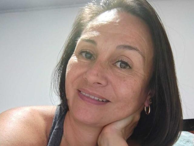 LatinLola has medium brown hair, a slight smile, and is resting her chin on her hand. She's wearing a gray top and small hoop earrings. Her tits are small.