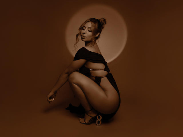 AmyBermudez squatting, wearing a black off-shoulder top, revealing her ass and back, with a side-tie thong, brown skin, and curly hair in a bun, against a brown backdrop.