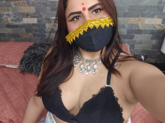 DaliaDubey with dark brown hair, wearing a black lace bra, yellow mask with gold trim, and red forehead dots, silver necklace, and black face mask, showing her tits.