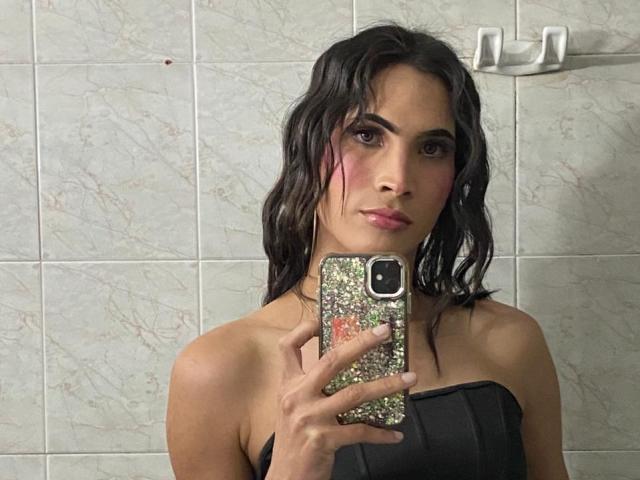 Alyice, con i capelli neri e bagnati, trucco rosa sugli occhi, che tiene un telefono scintillante, indossa un top nero senza spalline, in piedi in un bagno con piastrelle. Il seno è visibile, ma non si vedono glutei o genitali..