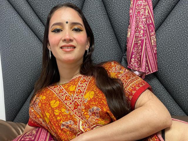 JalimaMona, with brown hair and a red and orange patterned sari, sits smiling, showing a bindi on her forehead, and pink earrings, with a pink sari hanging.